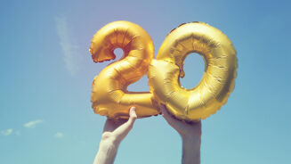 A gold foil number 20 balloon is held high in the air by caucasian male hand. The image has been taken outdoors on a bright sunny day, the sky is blue with some clouds. A vintage style effects has been added to the image.
