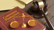 Golden scales of justice on a brown leather bound book engraved with the title Consumer Protection, together with a gavel and yellow office folders on a wooden table