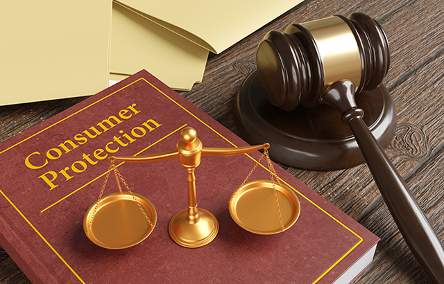 Golden scales of justice on a brown leather bound book engraved with the title Consumer Protection, together with a gavel and yellow office folders on a wooden table